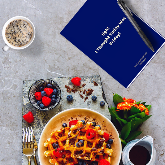 'Ugh! I Thought Today Was Friday!' 6X9 Navy blue lined notebook on a light grey marble table. A breakfast of waffles, syrup, berries and coffee is laid out and there are yellow/red flowers, on the side.